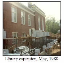 A brick library building under construction with scaffolding and materials around the foundation, trees in the background. Caption reads: Library expansion, May, 1980.