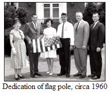 Six adults stand together outdoors holding a folded American flag. The image is black and white, with a caption reading “Dedication of flag pole, circa 1960.”.
