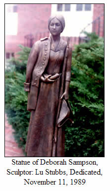 Bronze statue of Deborah Sampson wearing a long dress and holding a coat and hat, with greenery and a building in the background. Plaque notes sculptor Lu Stubbs and dedication date of November 11, 1989.