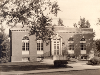 A sepia-toned photo of a small, brick building with three large arched windows and a central entrance, surrounded by trees and grass, with a sidewalk leading to the front steps.
